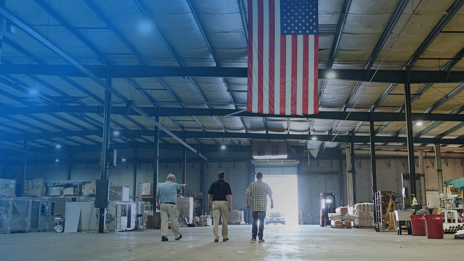 Three men walking through a large warehouse with an American flag hanging